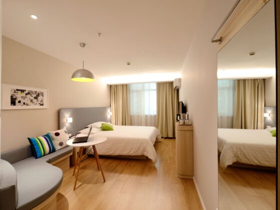 Hotel bedroom with a white bed, gray headboard, colorful pillows, a small round table with a laptop, and a gray sofa in a bright, modern space.