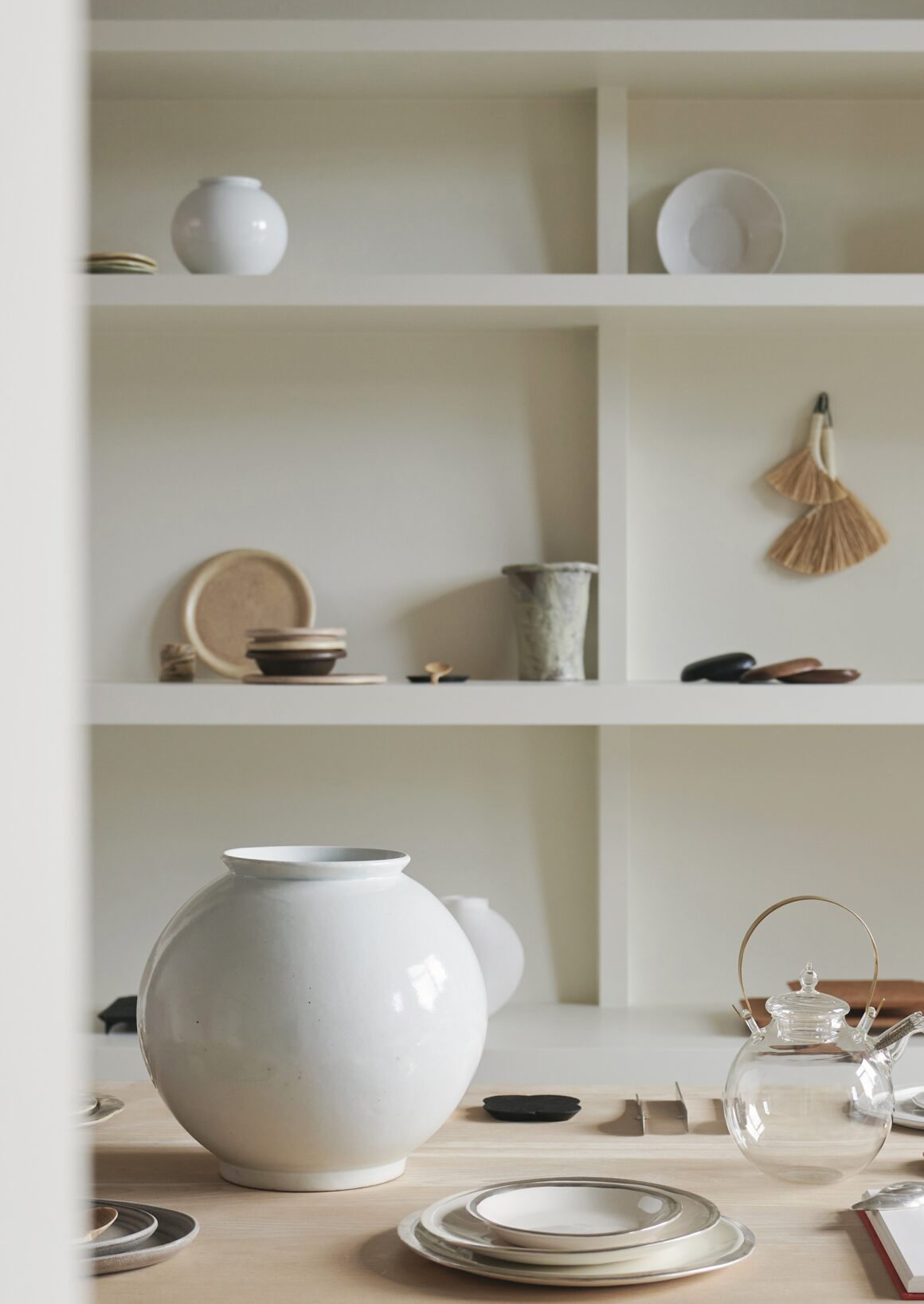 Open white shelf displaying ceramic dishes, bowls, and a large white vase with a hanging broom nearby.