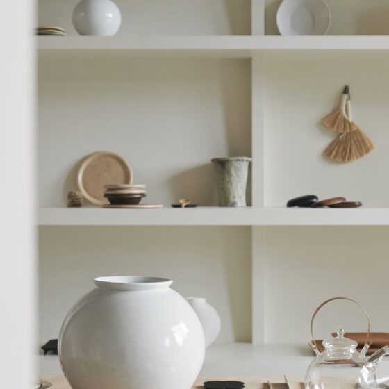 Open white shelf displaying ceramic dishes, bowls, and a large white vase with a hanging broom nearby.