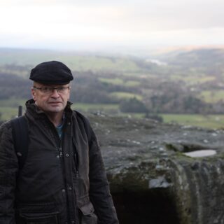Man wearing a black flat cap and brown jacket stands at a rocky overlook with a green valley and hills in the distance.