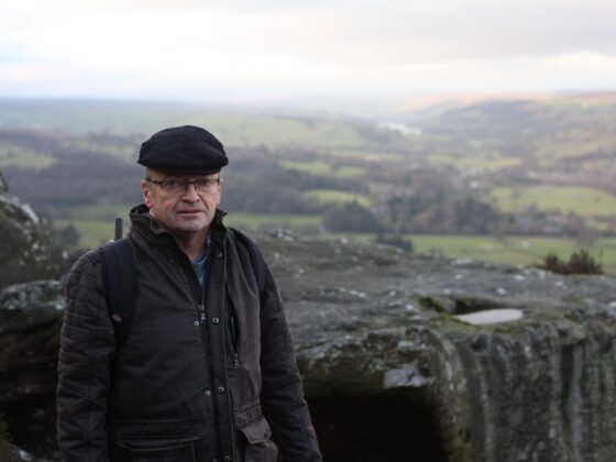 Man wearing a black flat cap and brown jacket stands at a rocky overlook with a green valley and hills in the distance.