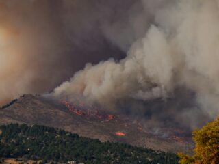 Hillside wildfire with flames and thick smoke rising over the ridge, with trees and homes at the bottom of the scene.