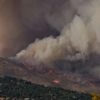 Hillside wildfire with flames and thick smoke rising over the ridge, with trees and homes at the bottom of the scene.