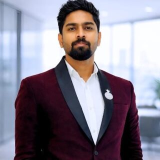 Professional man in a burgundy blazer with a circular badge, standing in a bright office space.