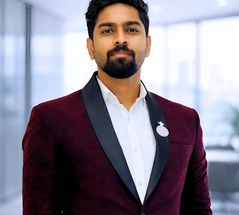 Professional man in a burgundy blazer with a circular badge, standing in a bright office space.