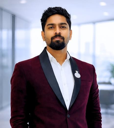 Professional man in a burgundy blazer with a circular badge, standing in a bright office space.