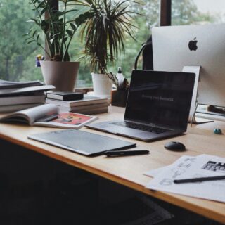 Desk setup with two potted plants, an open MacBook Pro showing a slide that reads 'Driving your business,' and a white iMac nearby with papers and books on the wooden desk.