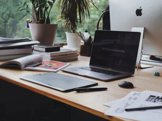 Desk setup with two potted plants, an open MacBook Pro showing a slide that reads 'Driving your business,' and a white iMac nearby with papers and books on the wooden desk.