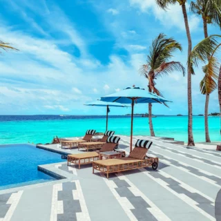 Resort poolside on a sunny beach with turquoise water, palm trees, blue umbrellas, and lounge chairs.