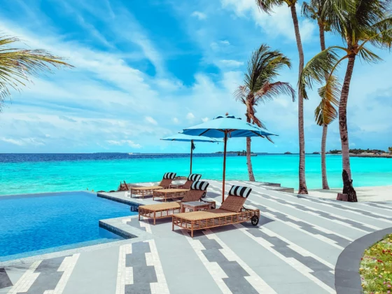 Resort poolside on a sunny beach with turquoise water, palm trees, blue umbrellas, and lounge chairs.