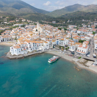 Aerial view of a coastal Mediterranean town with white buildings and terracotta roofs along a turquoise harbor, hillside in the background