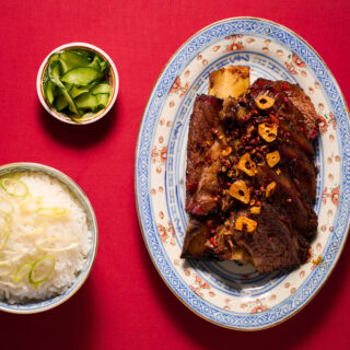 Plated braised beef with garlic chips and red pepper flakes on a decorative blue-and-white plate, with bowls of rice and cucumber salad on a red tablecloth.