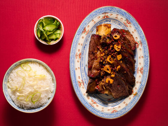 Plated braised beef with garlic chips and red pepper flakes on a decorative blue-and-white plate, with bowls of rice and cucumber salad on a red tablecloth.